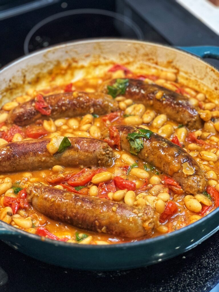 A pan of Italian sausage and beans. The hot sausage links are browned and laying in a pot of stewed tomatoes and white beans. The dish is garnished with fresh basil.
