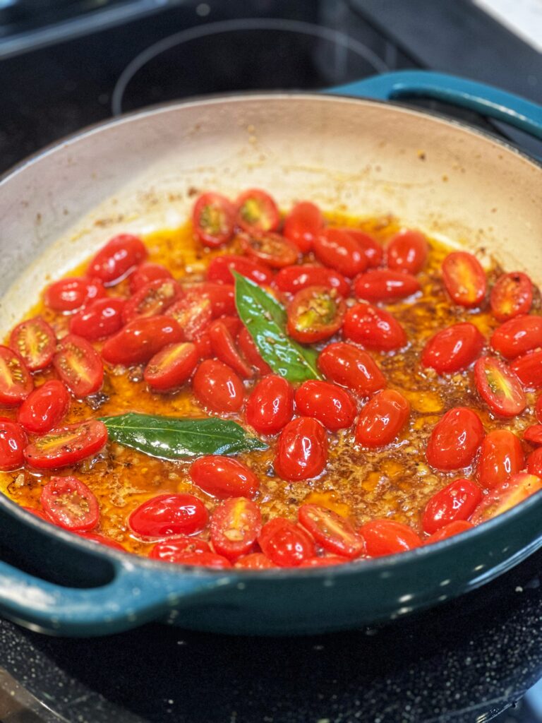 Halved cherry tomatoes simmering in a pan with minced garlic, bay leaves and chicken stock.