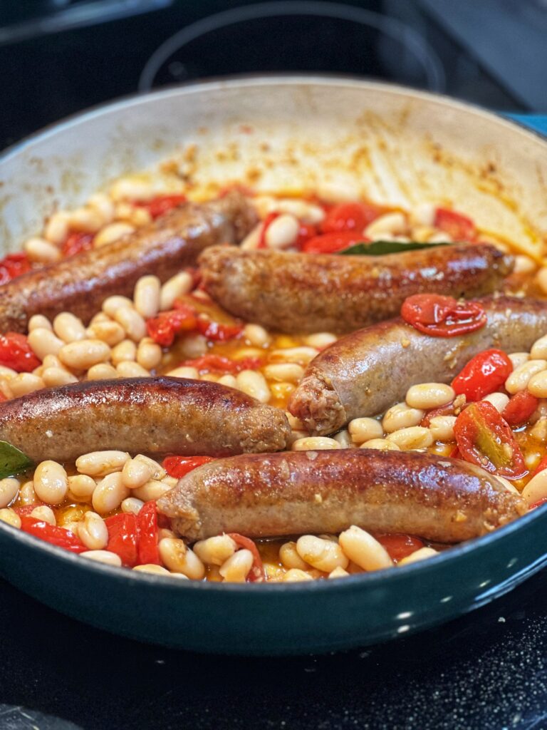 Browned sausage links in a pan with cooked cherry tomatoes and white beans.