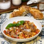 A white bowl of three browned hot sausage links in a stew of white beans and tomatoes, with fresh basil.