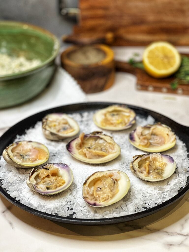 Eight shucked clams in their bottom shell laying in a flat cast iron pan that is lined with rock salt.