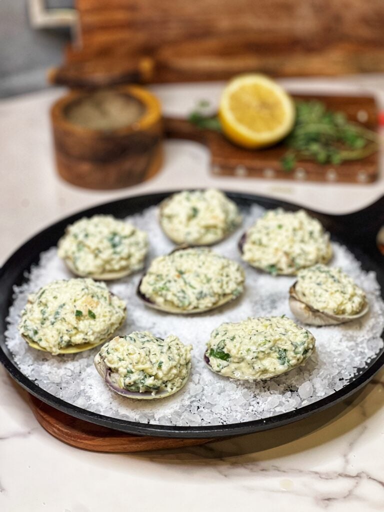 Eight shucked clams sitting in a flat cast iron pan lined with rock salt. The clams are filled with a creamy bread mixture mixed with fresh oregano.