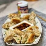 A white platter of shrimp and pork gyoza with a small rectangular bowl of soy sauce based dipping sauce in the middle of the platter.