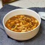 A bowl of hot and sour soup. The soup is brown with thin strips of pork, mushrooms, bamboo and tofu.