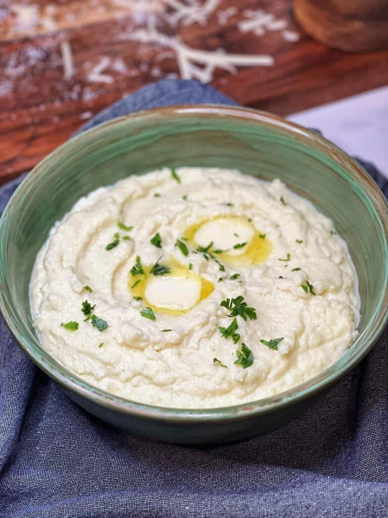 A deep green bowl filled with creamy cheesy mashed cauliflower that has been finished with a few pats of butter and chopped parsley.