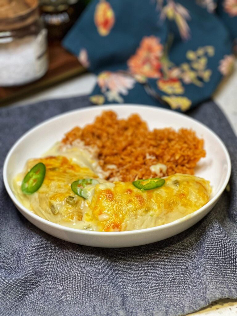 Two chicken enchiladas covered in sour cream sauce and topped with sliced jalapeños. It's served in a white bowl with Mexican rice.