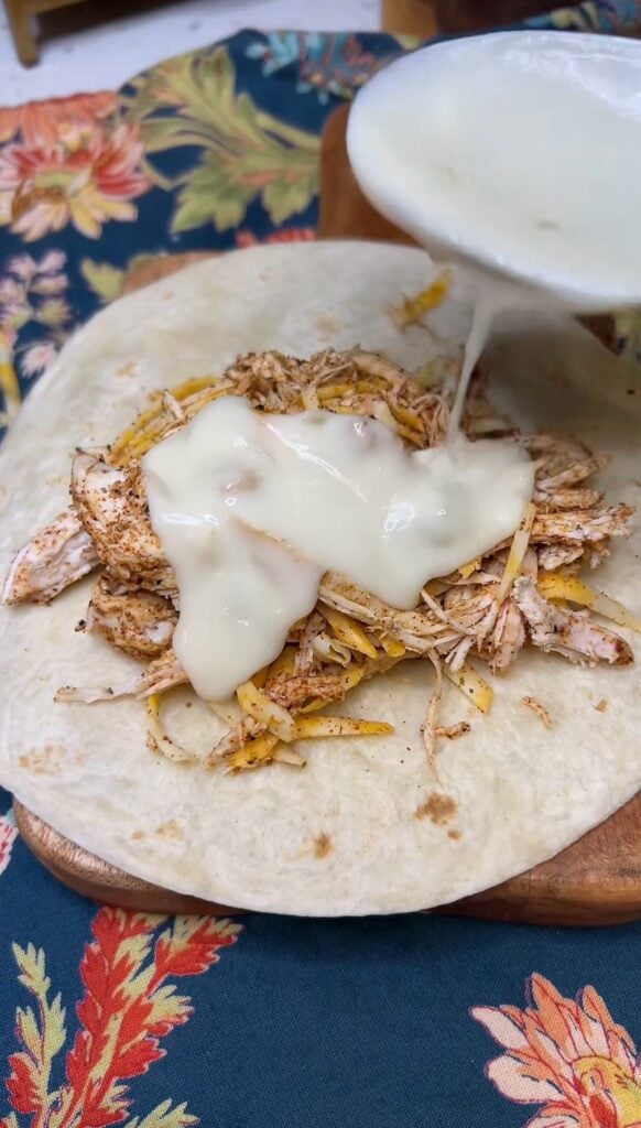 A laid our flour tortilla filled with seasoned shredded chicken with cheese. The photo shows topping with a spoonful of sour cream sauce.