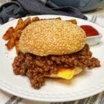 A sloppy joe sandwich sitting on a white plate with french fries and ketchup in the background. The sandwich is served on a sesame seed bun and has American cheese.