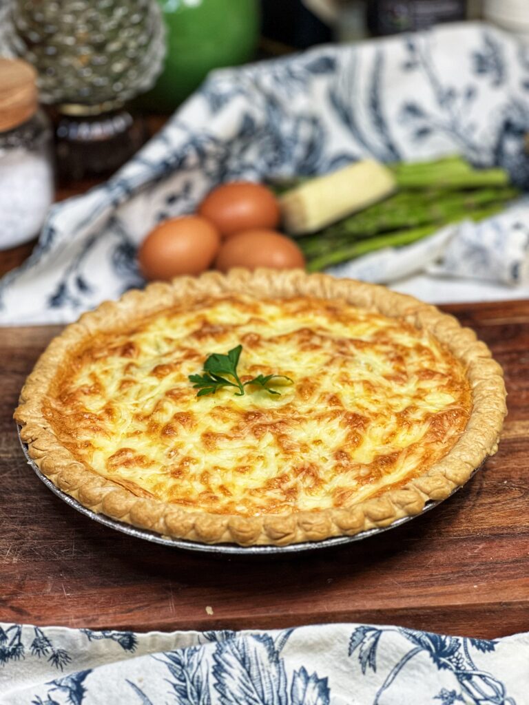 A deep-dish Asparagus Gruyère Quiche sitting on a wooden cutting board. The quiche has a golden brown crust and is garnished with a fresh parsley leaf.