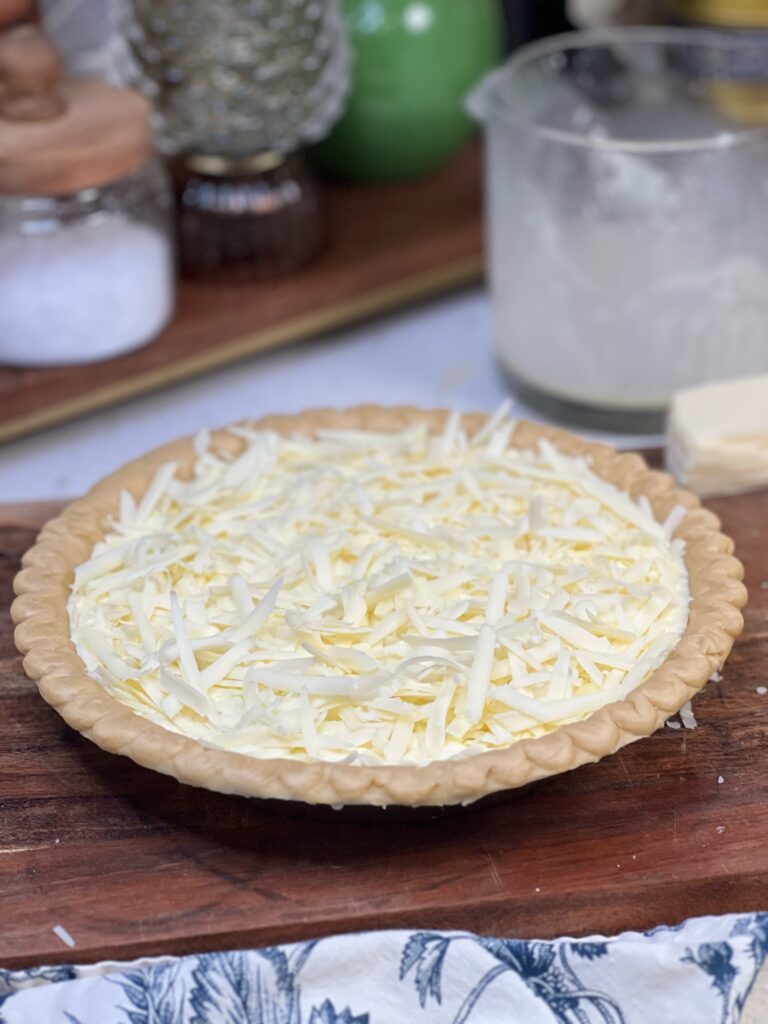 A frozen, deep-dish pie crust filled with an egg cream mixture and topped with shredded Gruyère cheese ready to be baked into an Asparagus Gruyère Quiche