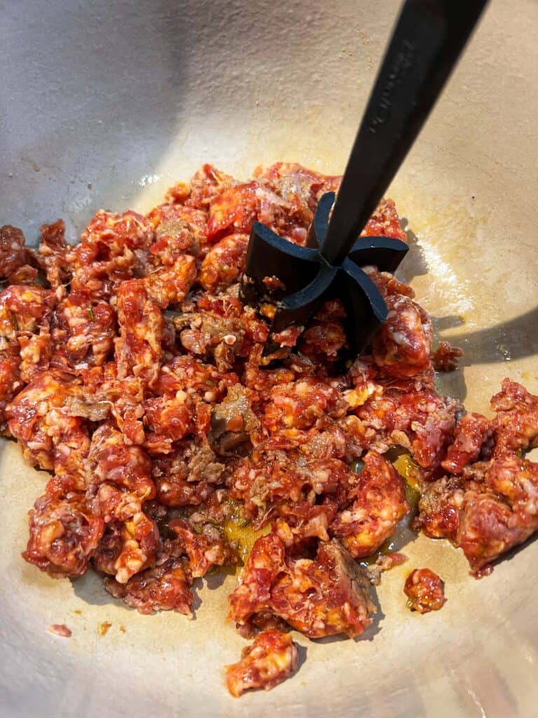 Using a meat chopper to break apart hot sausage in a pot.