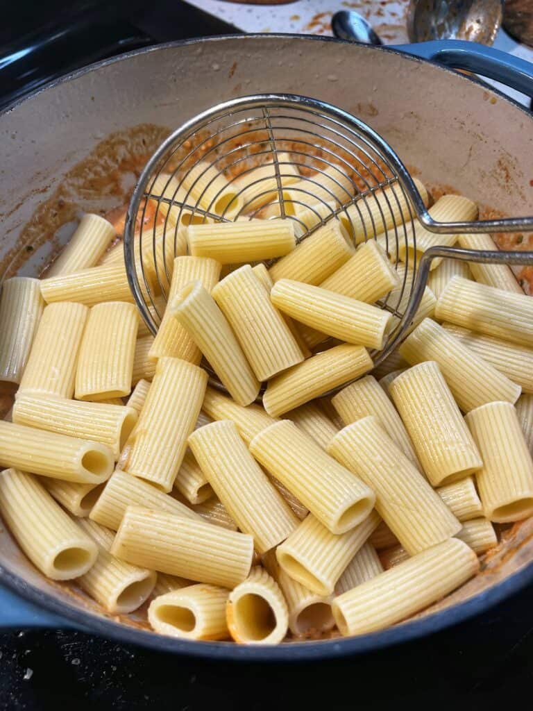 Cooked rigatoni being added with a slotted spoon into a pot of sausage cream sauce.