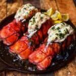 Three broiled lobster tails sitting on a flat cast iron pan. The tails are red in color. The lobster meat is sitting on top of the shells and garnished with fresh, chopped parsley.