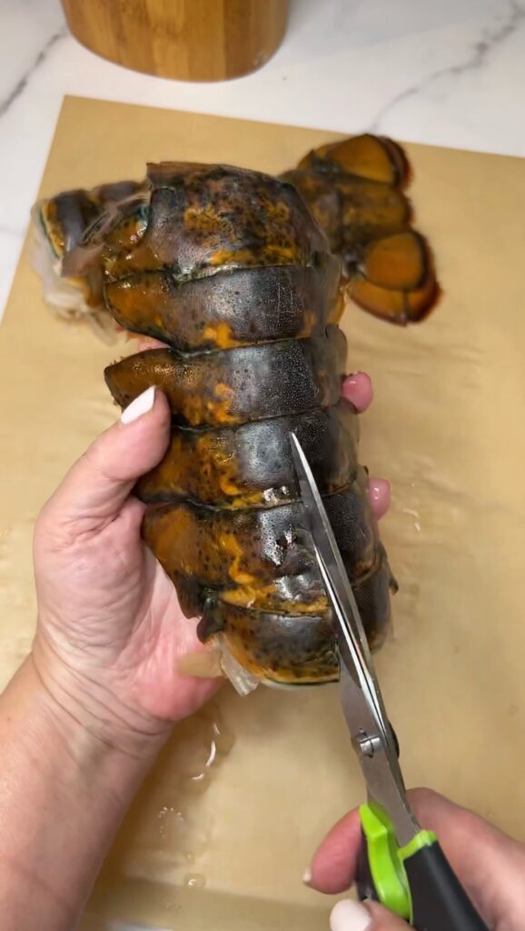 Using kitchen shears to cut the top shell in half of a raw lobster tail.