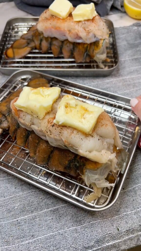 A raw lobster tail with the meat taken out of it's shell and laying on top of the shell. The meat is seasoned with salt, pepper, paprika with two butter pats on top for Broiled Lobster Tails.