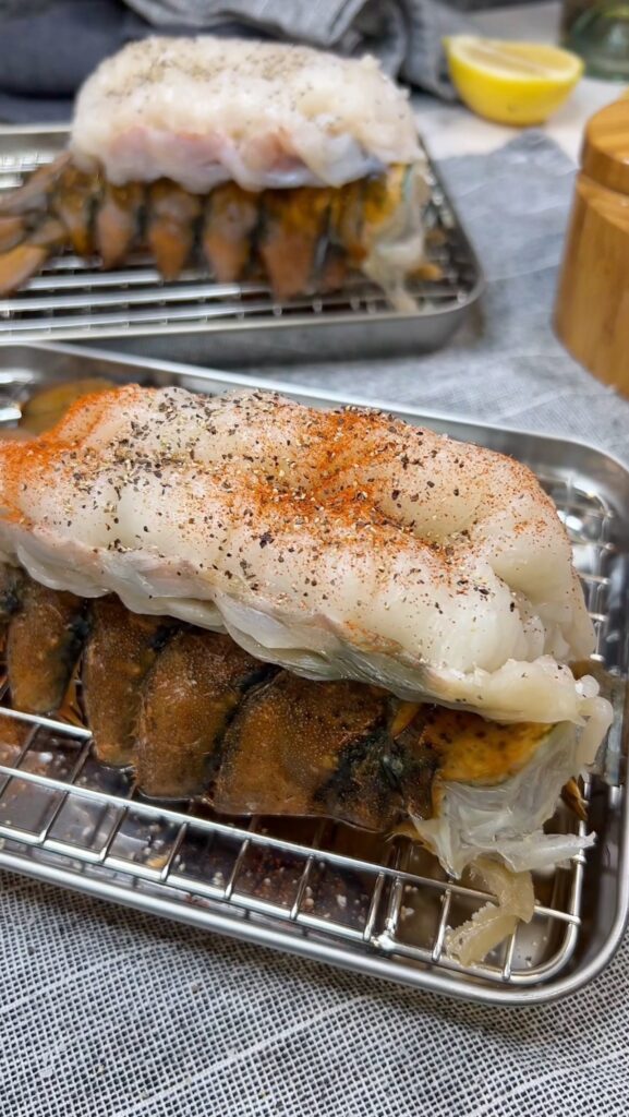 A raw lobster tail sitting on a small pan with a rack. The lobster meat is pulled out from the shell and laying on top of the shell. It's seasoned with salt, pepper and paprika.