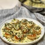 A white platter of seared scallops topped with a creamy lemon butter garlic sauce and garnished with fresh parsley.