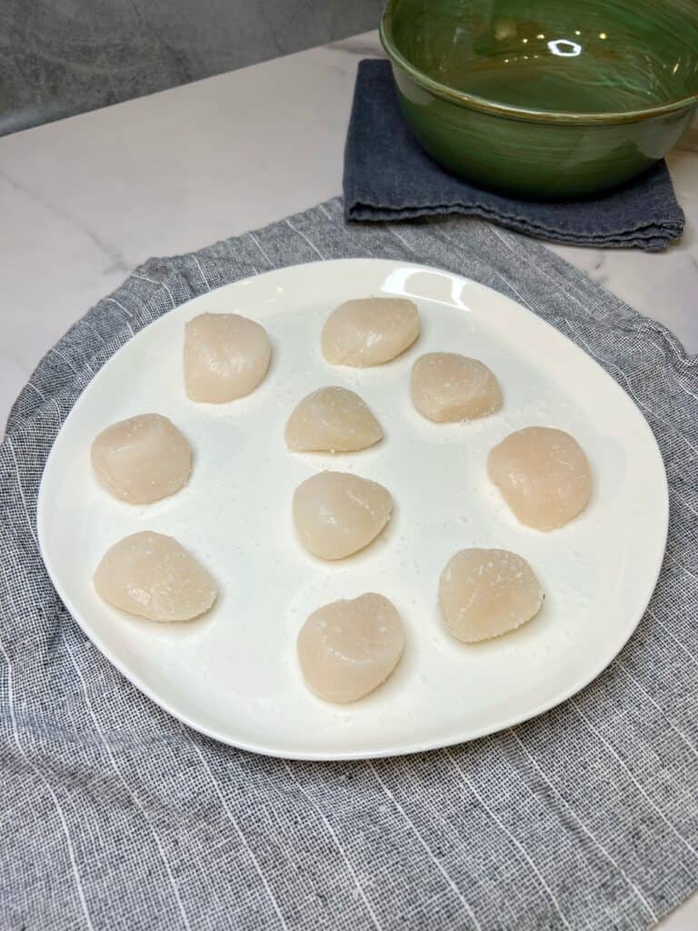 Ten scallops sitting on a white plate seasoned with salt.