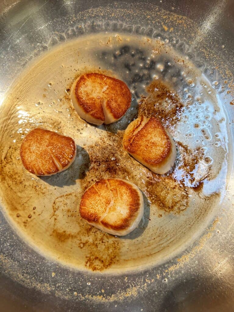 Four scallops searing in a stainless steel pan.