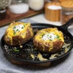 Two steakhouse baked potatoes sitting on a flat cast iron pan filled with melty cheese, sour cream and garnished with fresh parsley.