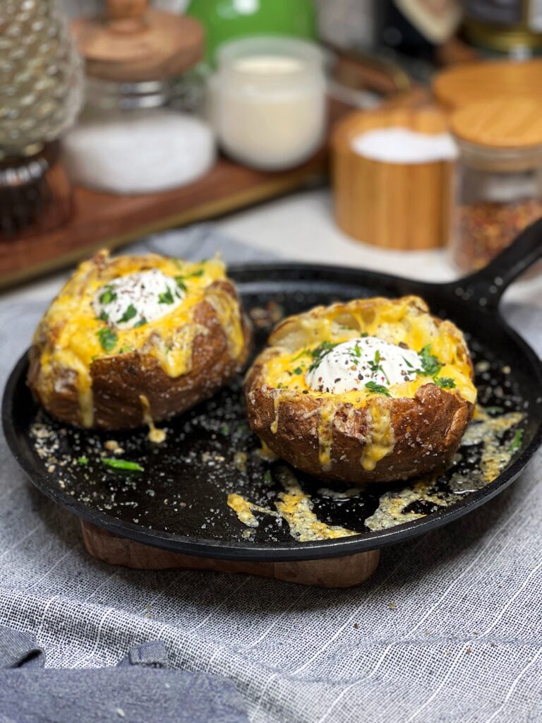 Two steakhouse baked potatoes sitting on a flat cast iron pan filled with melty cheese, sour cream and garnished with fresh parsley.