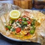 A piece of Mediterranean Baked Cod in parchment paper. The fish is seasoned and steamed in parchment with cherry tomatoes, white beans, olives and capers.