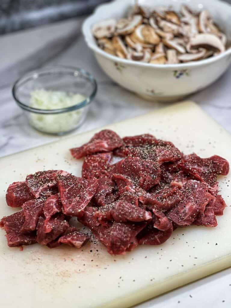 Thin sliced raw beef filet on a white plastic cutting board seasoned with salt and pepper for simple classic beef stroganoff.