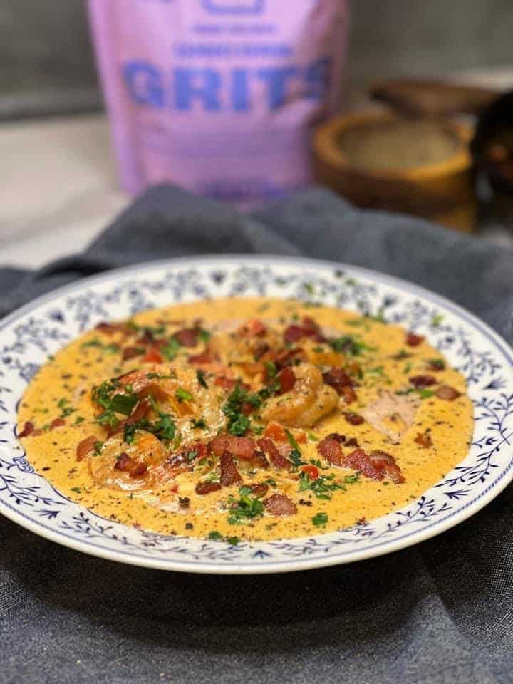 A wide white bowl trimmed in a blue decorative design filled with creamy cheesy shrimp and grits that are garnished with crisp bacon and fresh chopped parsley.
