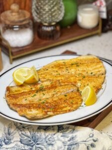 Two pieces of pan fried trout on a white platter garnished with fresh parsley and served with lemon wedges.