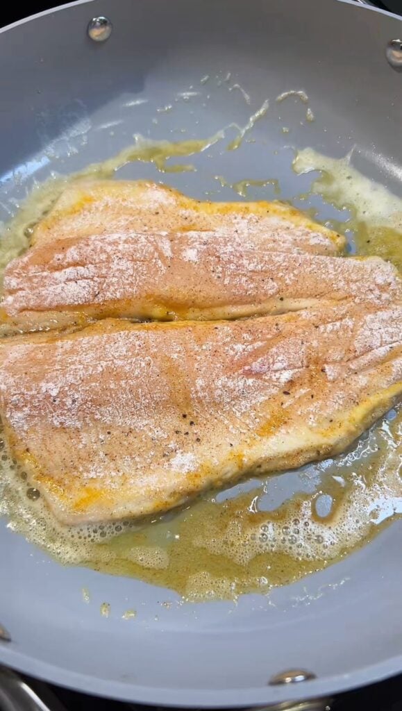 A piece of butterflied trout dredge in seasoned flour frying in a non-stick pan, skin side down.
