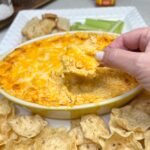 Me using a tortilla chip to scoop out Buffalo chicken dip from a casserole dish.