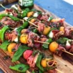 Prosciutto Melon Skewers with Basil Pesto on a wooden board. The skewers have cantaloupe balls, watermelon balls, mozzarella pearls, fresh basil and prosciutto.