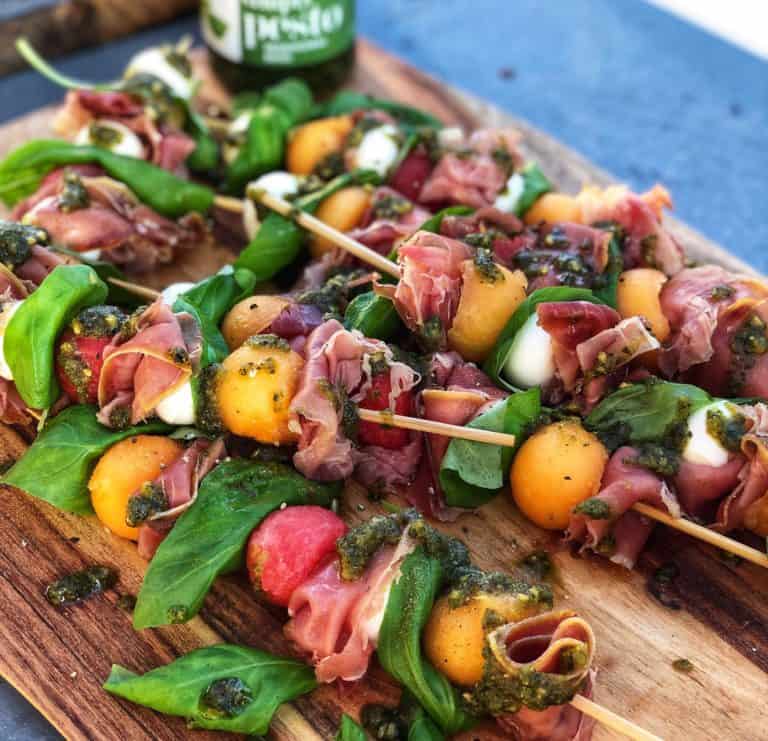 Prosciutto Melon Skewers with Basil Pesto on a wooden board. The skewers have cantaloupe balls, watermelon balls, mozzarella pearls, fresh basil and prosciutto.