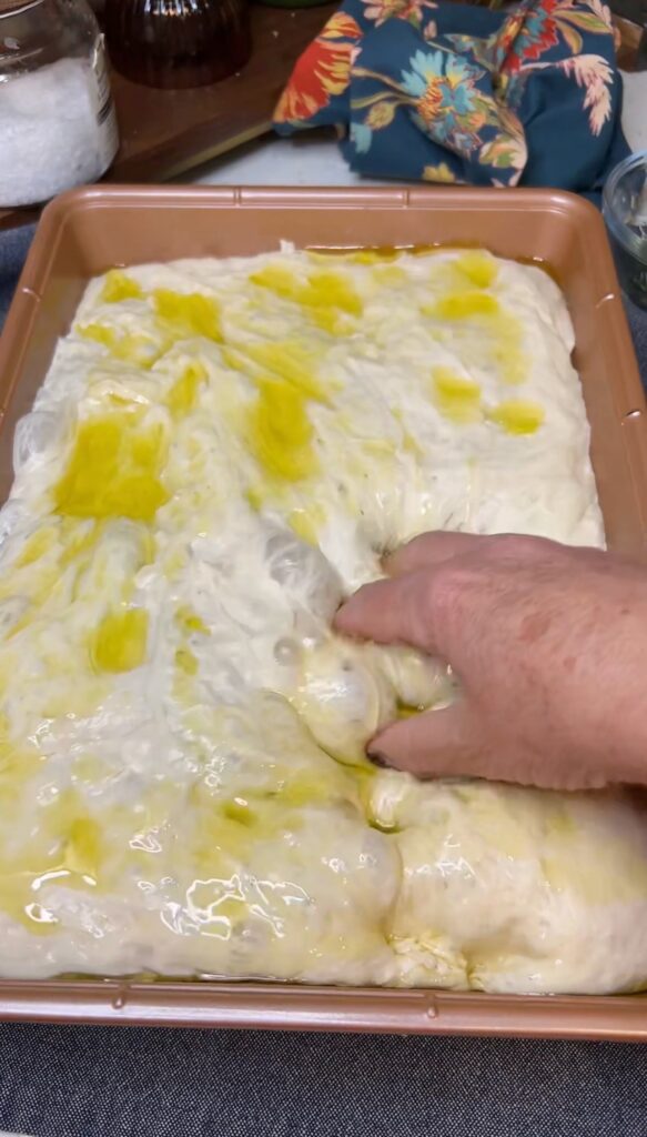 Focaccia dough in a pan coated with olive oil. I am dimpling the dough with my hand.