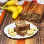 Slices of banana nut bread stacked on a plate with sliced bananas. Bananas and the loaf of banana bread is sitting in the background.