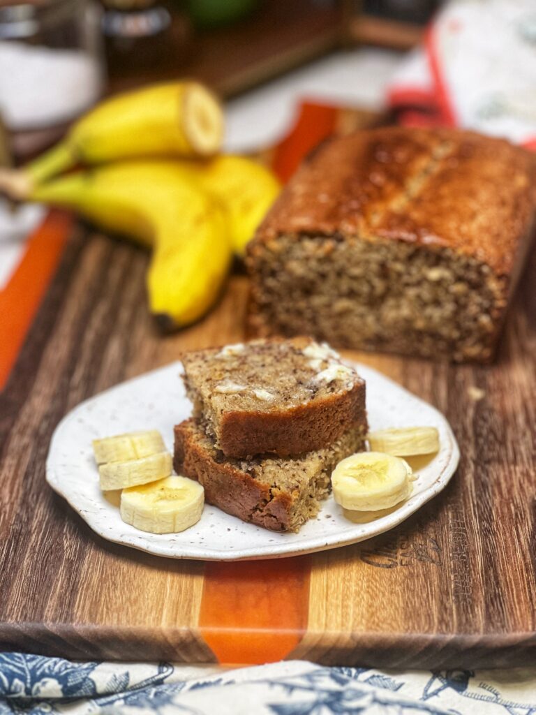 Slices of banana nut bread stacked on a plate with sliced bananas. Bananas and the loaf of banana bread is sitting in the background.