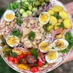 A classsic tuna niçoise salad on a platter. The salad has halved cherry tomatoes, canned tuna in the middle, hard boiled eggs, steamed potatoes, olives, cucumbers, green beans, white beans and red onion.