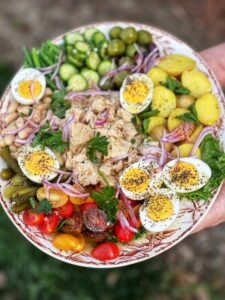 A classsic tuna niçoise salad on a platter. The salad has halved cherry tomatoes, canned tuna in the middle, hard boiled eggs, steamed potatoes, olives, cucumbers, green beans, white beans and red onion.