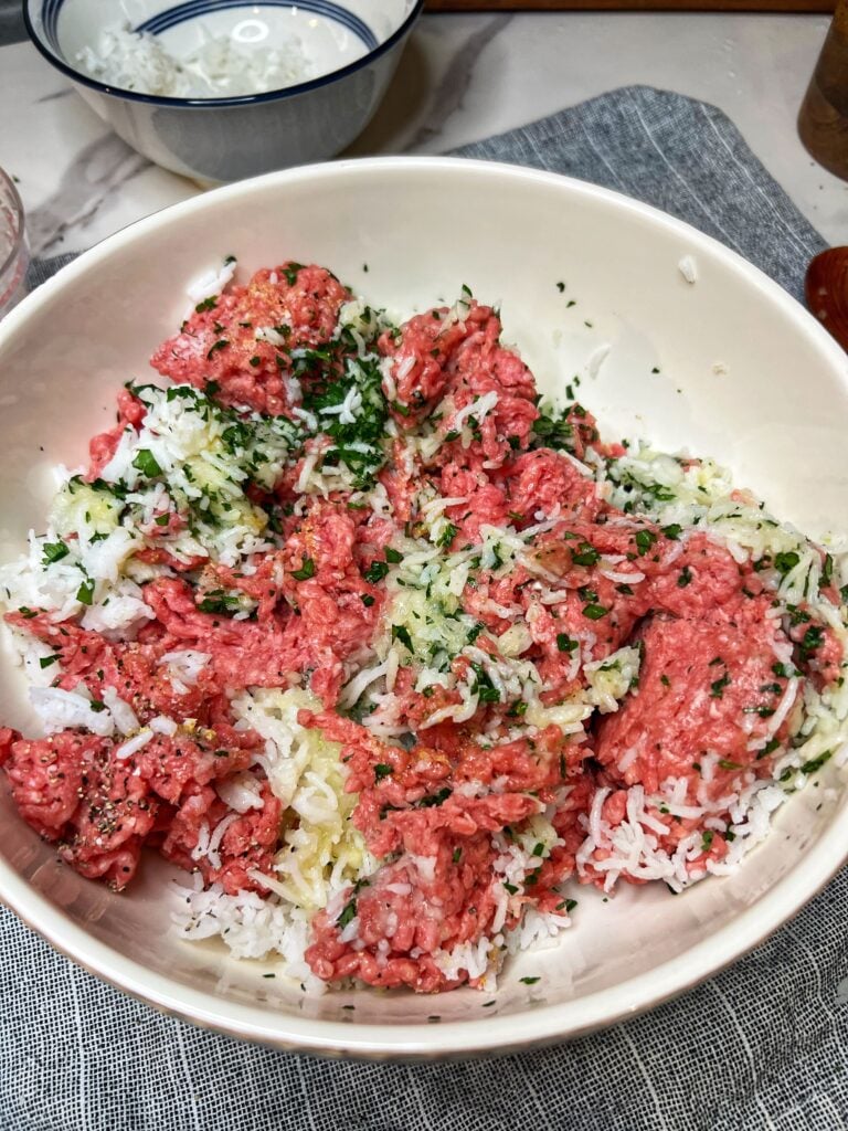 Ground beef, seasonings, rice, minced onions and fresh parsley in a large white bowl.