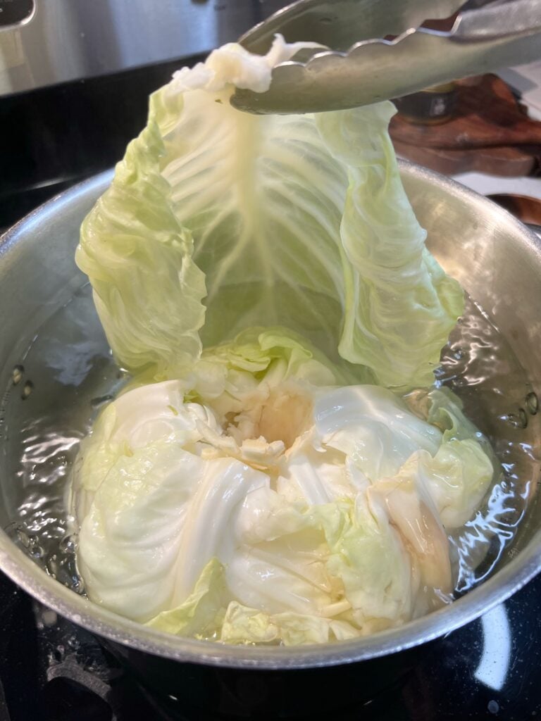 Removing a steamed cabbage leaf from a pot of simmering water with tongs.