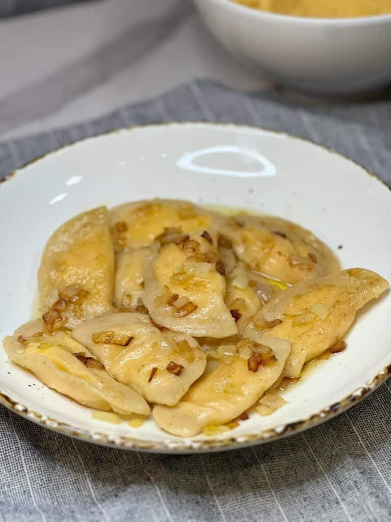 A gold rimmed white bowl filled with homemade potato and cheese pierogies covered in brown butter and onions.