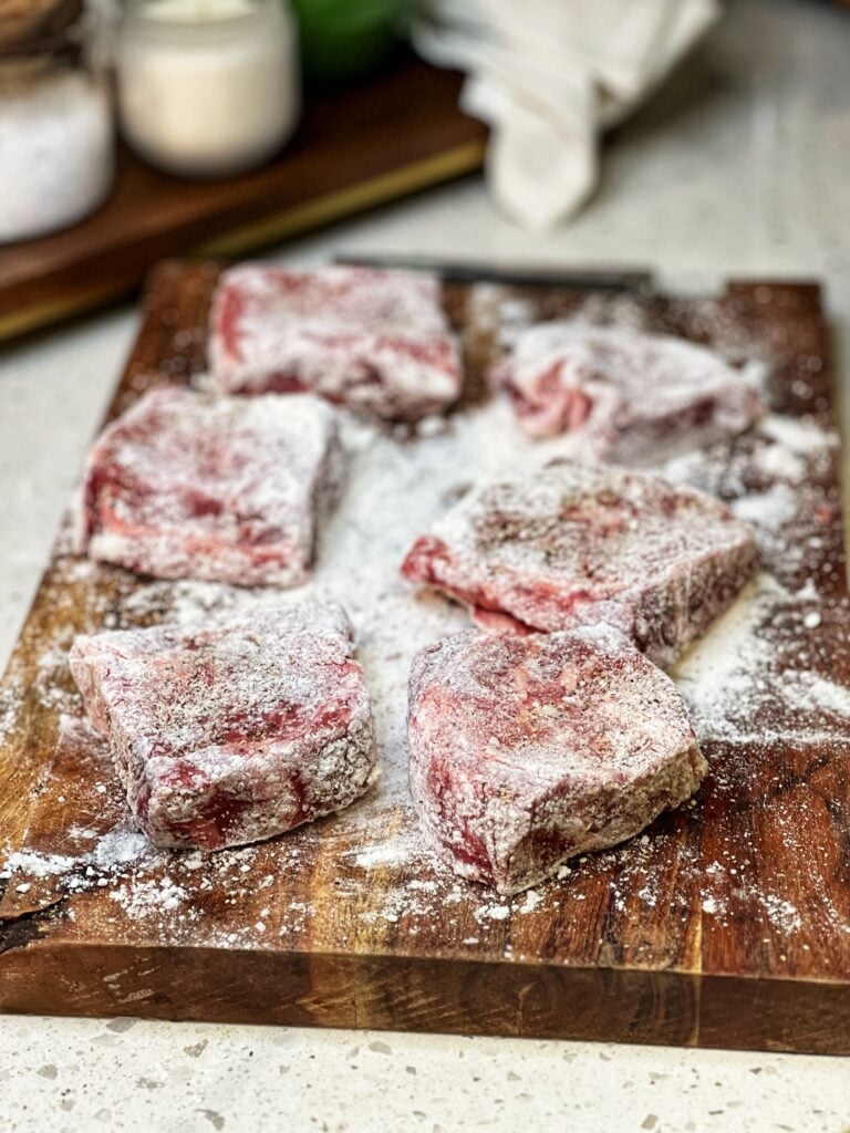Six pieces of steak coated in flour sitting on a wooden board.