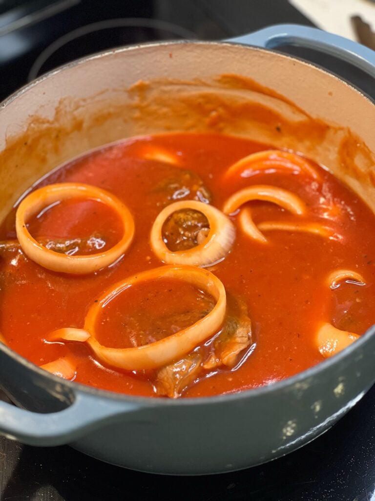 A deep pot filled with seared steak and thick-sliced white onion in a red gravy.