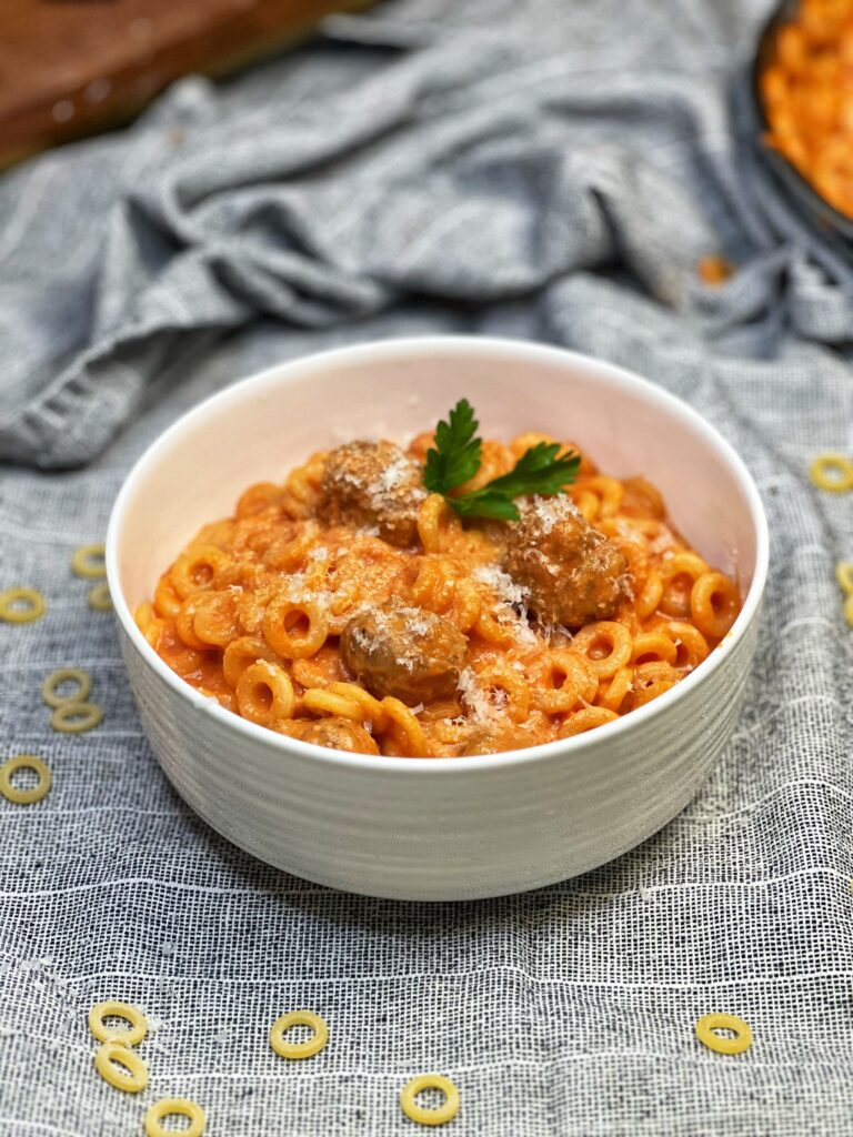 A white bowl of Spaghetti O's Pasta with Meatballs. The dish is garnished with fresh parsley and freshly grated Parmesan cheese.