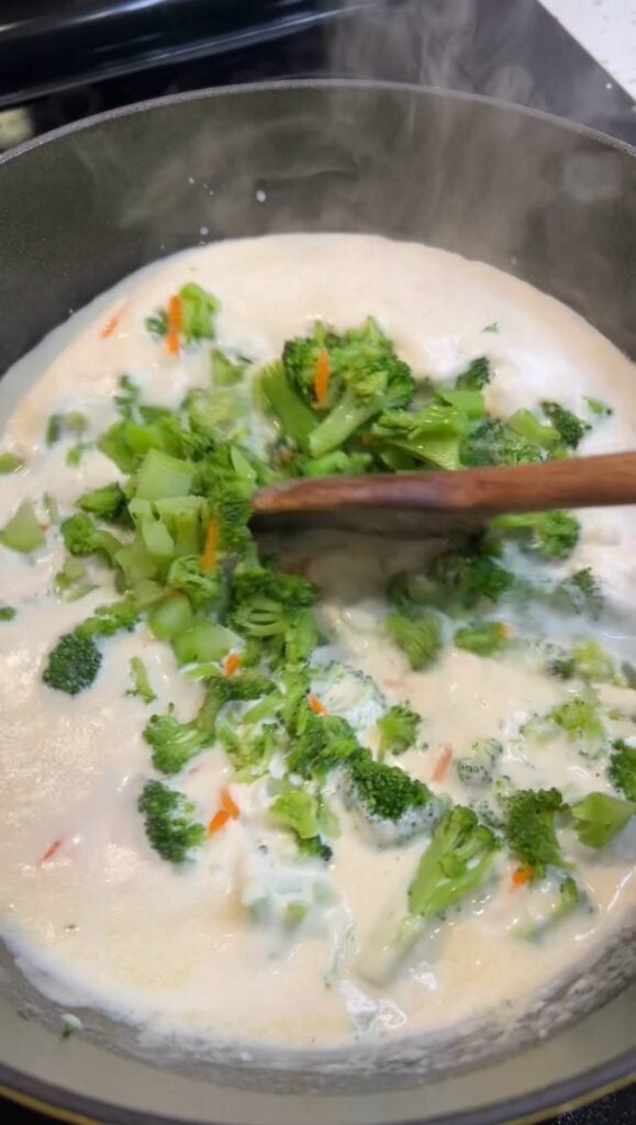 A pot of a creamy soup base with chopped broccoli and carrots being stirred in for Broccoli Cheddar Cheese Soup.