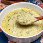 A white bowl of Broccoli Cheddar Cheese Soup with a wooden spoonful of the soup being shown over the bowl.
