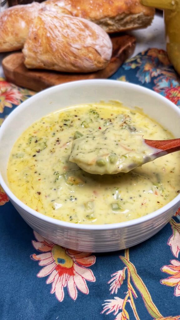 A white bowl of Broccoli Cheddar Cheese Soup with a wooden spoonful of the soup being shown over the bowl.