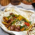 A piece of red snapper that has been steamed in parchment paper with halved cherry tomatoes and snow peas. Roasted potatoes have been placed inside the opened parchment packet with the fish.
