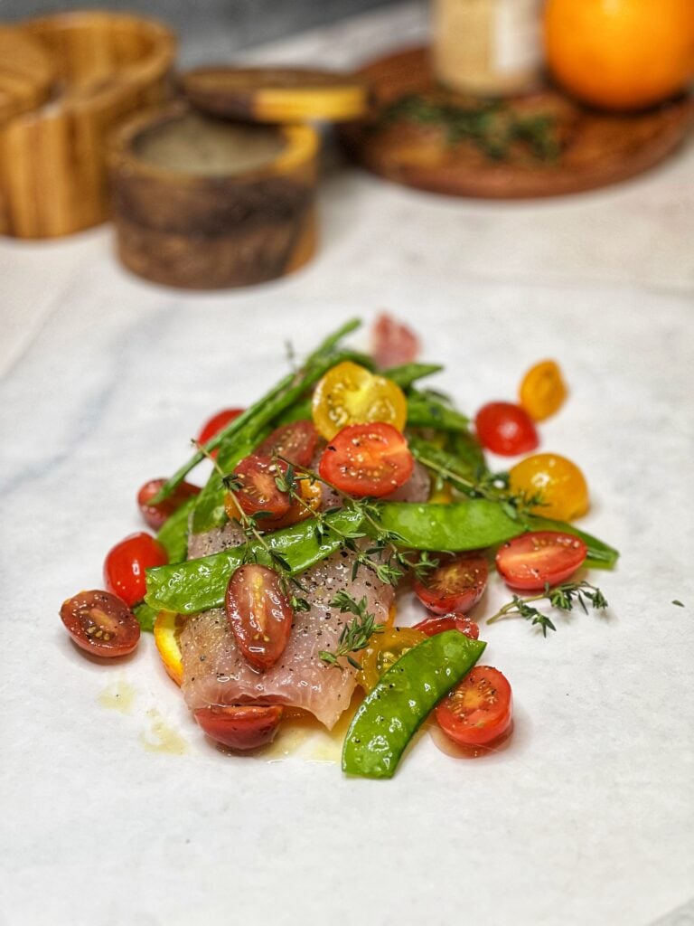 A raw, red snapper filet sitting on top of orange and lemon slices that have been laid on top of a piece of white parchment paper. This fish is topped with halved cherry tomatoes, snow peas and fresh herbs for a recipe for Baked Fish in Parchment.