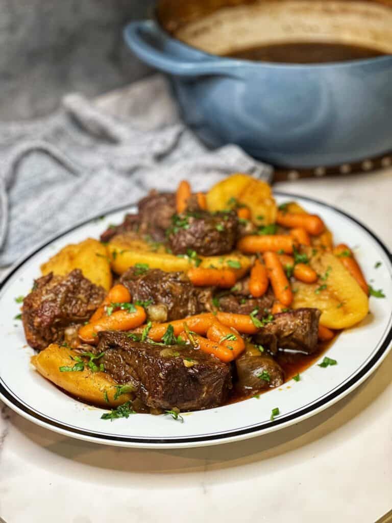 An oval white platter trimmed in black holding beef roast chunks with cooked potatoes and carrots. Everything is garnished with fresh parsley.